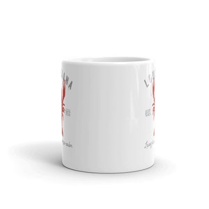 May include: A white ceramic mug featuring a red crawfish graphic centered between the text LOUISIANA and EST. 1812 in grey lettering. Below the crawfish, the cursive text Laissez les bons temps rouler is printed in grey on this cylindrical drinkware.