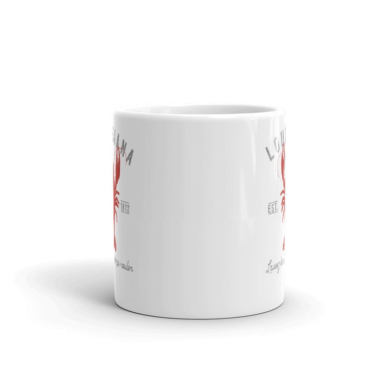 May include: A white ceramic mug featuring a red crawfish graphic centered between the text LOUISIANA and EST. 1812 in grey lettering. Below the crawfish, the cursive text Laissez les bons temps rouler is printed in grey on this cylindrical drinkware.