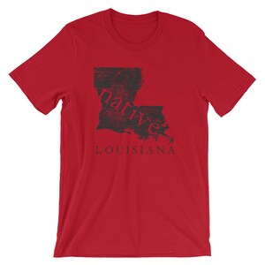 May include: A red short-sleeved t-shirt featuring a black graphic of the state of Louisiana. The word native is printed in red across the center of the state map, and the word LOUISIANA is printed in black capital letters below the map graphic.
