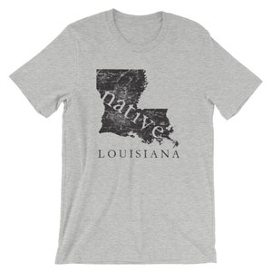 May include: A heather grey short-sleeve t-shirt featuring a black, distressed-texture graphic of the state of Louisiana. The word "native" is printed in white lowercase letters across the map, with the word "LOUISIANA" in black below it.