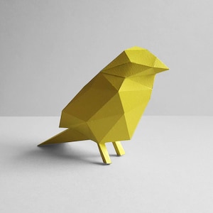 Può includere: Una scultura di uccello di carta gialla con un design geometrico. L'uccello è in piedi su una superficie bianca.