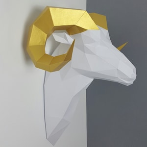 May include: A white papercraft ram head with gold horns. The ram's head is a 3D paper model with a geometric design.