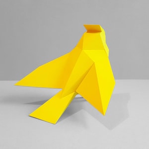 Può includere: Una scultura di uccello origami gialla con un design geometrico. L'uccello è in piedi su una superficie grigia.