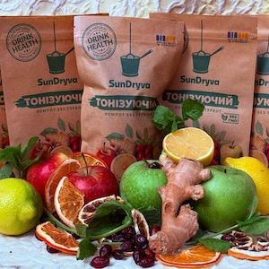 Puede incluir: Bolsas de papel marrón con el texto "SunDryva" y "Drink Health" se muestran con fruta fresca y jengibre. Las bolsas están rodeadas de manzanas, limones, naranjas y arándanos, lo que sugiere un producto saludable.