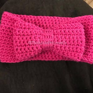 Puede incluir: Una diadema de ganchillo fucsia vibrante. La diadema presenta un patrón de puntada texturizada y un detalle de lazo anudado en el centro. El texto "@ThisLittlePrincessCo" es visible en la diadema.