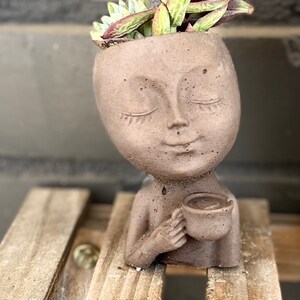 May include: A brown ceramic planter shaped like a person with closed eyes and a smile, holding a small cup. The planter is filled with a variety of succulents, including green and red varieties.