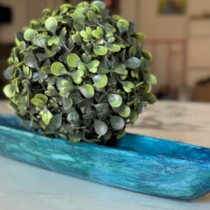 May include: A green artificial plant in a blue and green wooden bowl. The bowl is shaped like a boat.