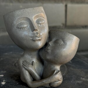 May include: A gray ceramic sculpture of two faces embracing. The faces are stylized and have closed eyes. The sculpture is a planter and has a hole in the top of each head.