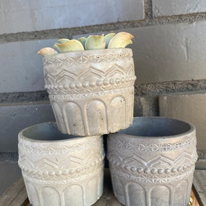 May include: Three gray concrete planters with a geometric design. The top planter has a small succulent plant inside.
