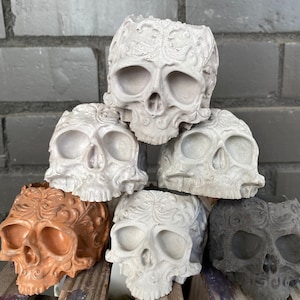 May include: Six decorative skull planters made of concrete. The planters are stacked in a pyramid shape. The top planter is white, the next two are white with intricate designs, the next two are white with brown designs, and the bottom planter is gray.