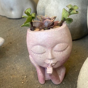 May include: A pink concrete planter shaped like a face with closed eyes and a finger to its lips. The planter is filled with a succulent plant with green leaves.