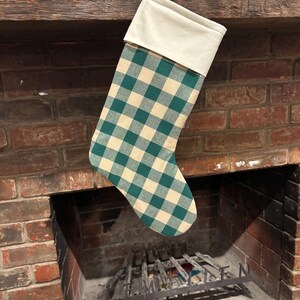 May include: A Christmas stocking featuring a green and cream-colored buffalo check pattern on the main body and a solid cream-colored cuff at the top. The stocking includes a small fabric loop attached to the upper corner for hanging purposes.