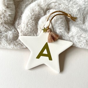 Peut inclure: Une étoile en céramique blanche avec une lettre A dorée et un pompon brun. L'étoile a un charme d'étoile dorée attaché au pompon.