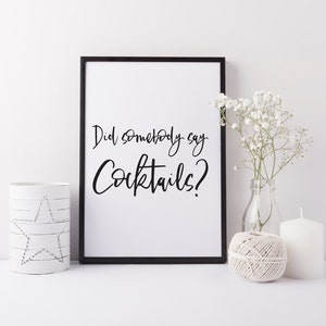 K&ouml;nnte beinhalten: Schwarz-wei&szlig;es druckbares Wandbild mit dem Text "Did somebody say Cocktails?" in einer handschriftlichen Schriftart. Der Druck ist in einem schwarzen Rahmen gerahmt und wird auf einer wei&szlig;en Oberfl&auml;che mit wei&szlig;en Blumen und Kerzen pr&auml;sentiert.