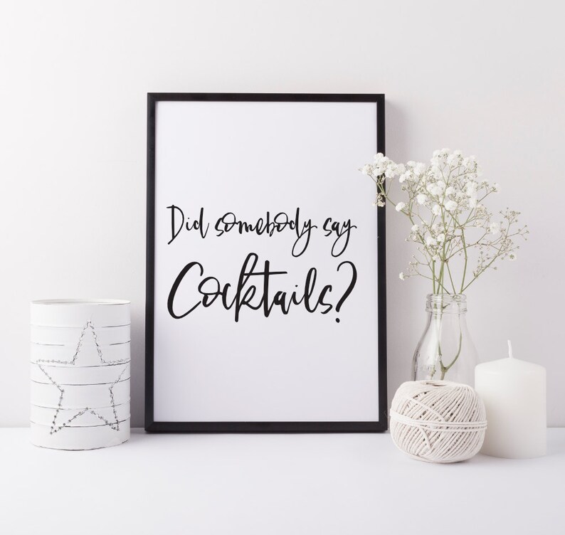K&ouml;nnte beinhalten: Schwarz-wei&szlig;es druckbares Wandbild mit dem Text "Did somebody say Cocktails?" in einer handschriftlichen Schriftart. Der Druck ist in einem schwarzen Rahmen gerahmt und wird auf einer wei&szlig;en Oberfl&auml;che mit wei&szlig;en Blumen und Kerzen pr&auml;sentiert.