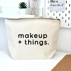 May include: A white canvas makeup bag with a zipper closure. The bag has black text that reads "makeup + things."