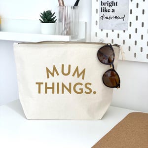 May include: A white canvas zippered pouch with the text "MUM THINGS." printed in gold. The pouch is lying on a cork surface with a pair of brown sunglasses hanging from the zipper.