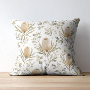 May include: Square cushion with a botanical pattern. The cushion features a repeating design of beige banksia flowers and green leaves on a white background. The cushion is resting on a wooden surface.