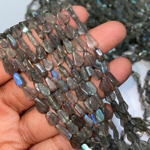 Op de afbeelding: Strengen grijze labradoriet edelsteen kralen. De kralen zijn druppelvormig en aan elkaar geregen. Sommige kralen hebben iriserende blauwe en groene flitsen. De kralen worden in een hand tegen een witte achtergrond gehouden.