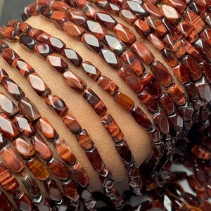 Puede incluir: Primer plano de múltiples hilos de pulseras de piedras preciosas de ojo de tigre rojo. Las cuentas son una mezcla de tonos marrón rojizo, con una forma pulida y facetada. Las pulseras están apiladas en una muñeca, mostrando las variaciones naturales de las piedras.
