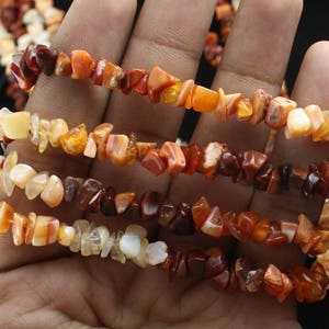Op de afbeelding: Close-up van verschillende armbanden gemaakt van kleine, onregelmatig gevormde, gepolijste edelstenen. De stenen zijn in tinten oranje, bruin en crème. De armbanden worden in een hand gehouden tegen een donkere achtergrond.