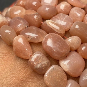 May include: A strand of sunstone beads, featuring a mix of smooth, polished, and tumbled stones. The beads are a light peach color with a subtle shimmer.