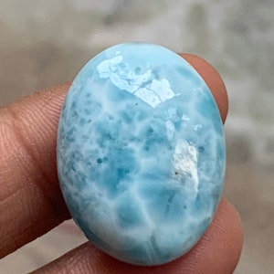 May include: Oval-shaped, polished gemstone in shades of light blue and white, resembling ocean waves. The stone has a smooth, glossy surface and is held between fingers. The gemstone is a Larimar.