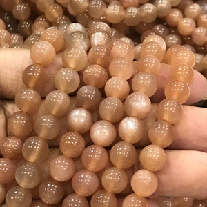 May include: Close-up of strands of peach-colored, round gemstone beads. The beads have a translucent quality, with a subtle sheen and variations in color. The beads are held in a hand, suggesting they are ready to be used for jewelry making.