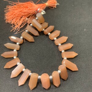 Op de afbeelding: Een streng peach-kleurige sunstone kralen met zilveren tussenstukken. De kralen zijn facetgeslepen en hebben een puntige vorm. De streng is afgewerkt met een kwast.