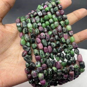 May include: A strand of tumbled ruby zoisite beads featuring a mix of vibrant magenta, deep forest green, and black mineral inclusions. The polished, irregular pebble-shaped stones are strung together in a multi-row arrangement with smooth surfaces.