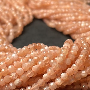 May include: A strand of peach-colored sunstone beads, faceted and polished, with a shimmery, iridescent finish. The beads are small and round, perfect for jewelry making.