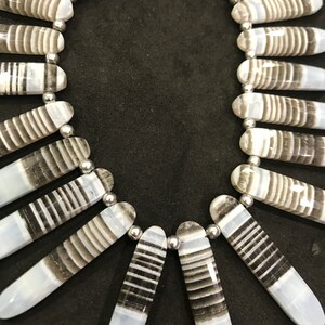 Peut inclure: Un collier composé de perles d'agate rayées de blanc et de gris avec des entretoises argentées. Les perles sont longues et étroites, et elles sont disposées en forme d'éventail.