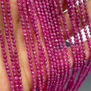 May include: Multiple strands of small, round, faceted magenta-pink gemstone beads. Each bead features a reflective, multi-faceted surface that catches the light, creating a uniform, vibrant appearance across the long, thin, vertical strings.
