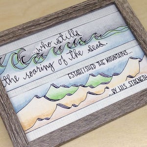 May include: Framed watercolor art print with a gray wooden frame. The artwork features hand-lettered text and watercolor illustrations of ocean waves and mountains. The text reads "...who stills the roaring of the seas. ESTABLISHED THE MOUNTAINS...BY HIS STRENGTH."