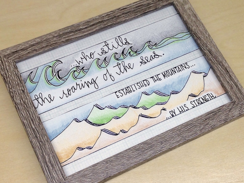 May include: Framed watercolor art print with a gray wooden frame. The artwork features hand-lettered text and watercolor illustrations of ocean waves and mountains. The text reads "...who stills the roaring of the seas. ESTABLISHED THE MOUNTAINS...BY HIS STRENGTH."