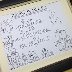 May include: Framed art print with black ink illustrations and handwritten text. The artwork includes floral and diamond shapes, and the words "STANING IN AWE P.2" and "THE pastures of the wilderness overflow". The frame is black with a gold inner border.