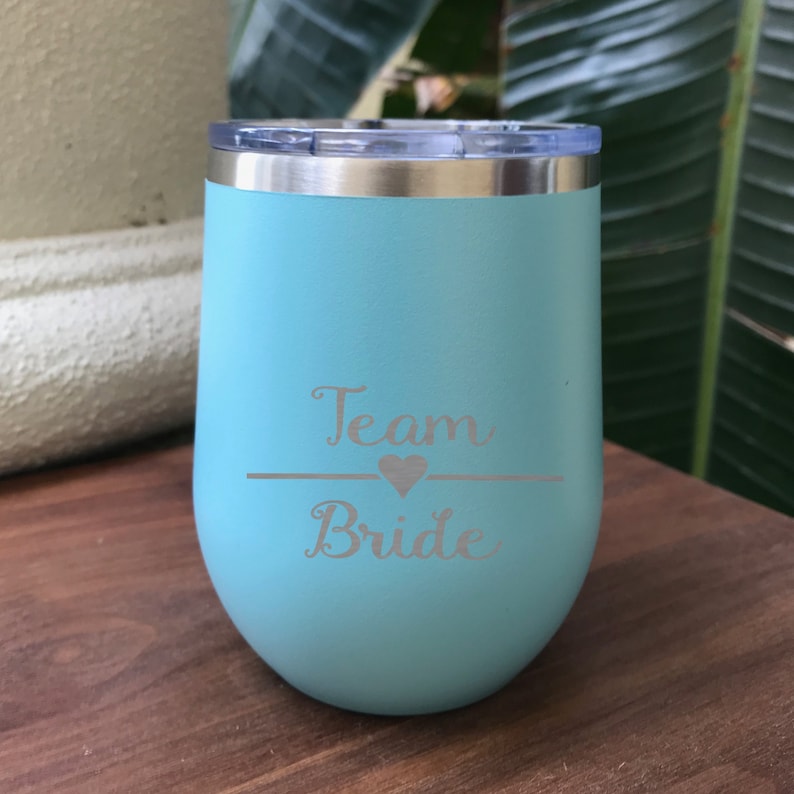 May include: A light blue stainless steel tumbler with a lid. The tumbler has the words "Team Bride" engraved on it with a heart symbol.