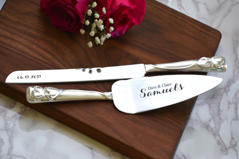 May include: Silver cake knife and server set with engraved hearts on the handles. The server is engraved with "Dave & Claire Samuels". The knife is engraved with "06 17 2017".