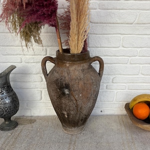 Può includere: Una natura morta con una brocca decorativa in metallo, un grande vaso marrone con manici e una ciotola di legno con frutta. Il vaso contiene fiori secchi. La frutta comprende mele, arance e una banana.