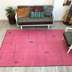 K&ouml;nnte beinhalten: Ein rosafarbener Teppich mit einem geometrischen Muster aus schwarzen Linien und Pfeilen. Der Teppich liegt auf einem Holzboden in einem Raum mit einer wei&szlig;en Couch und einem Stuhl.