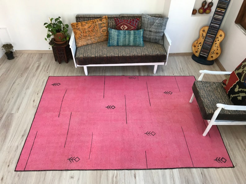 K&ouml;nnte beinhalten: Ein rosafarbener Teppich mit einem geometrischen Muster aus schwarzen Linien und Pfeilen. Der Teppich liegt auf einem Holzboden in einem Raum mit einer wei&szlig;en Couch und einem Stuhl.