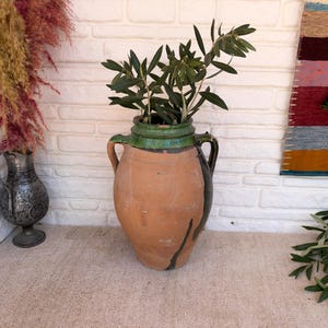 Pode incluir: Uma grande ânfora de terracota com duas alças, apresentando uma borda esmaltada em verde e uma faixa vertical verde-escura em um dos lados. O vaso contém ramos de oliveira. À esquerda, encontra-se um jarro metálico escuro menor.