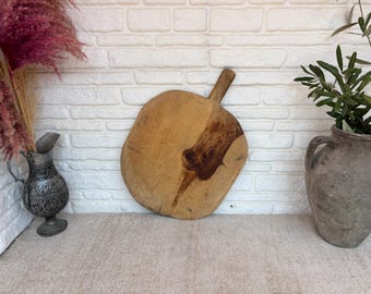 May include: A round wooden cutting board with a short handle, featuring a prominent dark brown wood grain knot in the center. The board has a light tan natural finish and a smooth, flat surface, showcasing the organic texture of the wood grain.