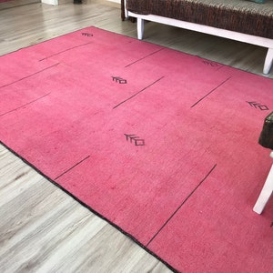 K&ouml;nnte beinhalten: Ein rosa Teppich mit einem geometrischen Muster in Schwarz. Der Teppich hat einen schwarzen Rand und liegt auf einem Holzboden.