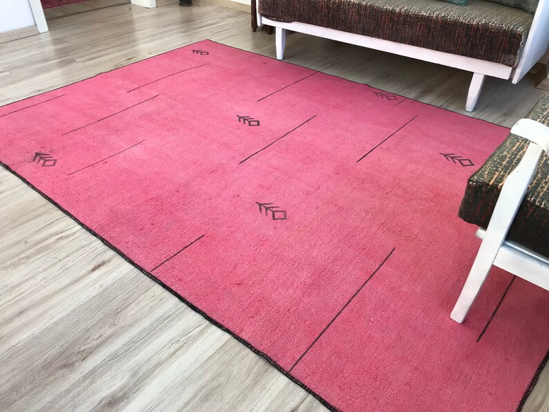 K&ouml;nnte beinhalten: Ein rosa Teppich mit einem geometrischen Muster in Schwarz. Der Teppich hat einen schwarzen Rand und liegt auf einem Holzboden.