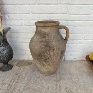 May include: A large, weathered, brown clay jug with a rounded body, a wide mouth, and a single side handle. The surface has a textured, mottled appearance with visible signs of age and wear, showcasing a rustic, earthy aesthetic.