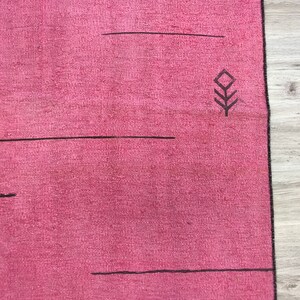 K&ouml;nnte beinhalten: Ein rosa Teppich mit einem schwarzen geometrischen Muster. Der Teppich hat einen Fransenrand.