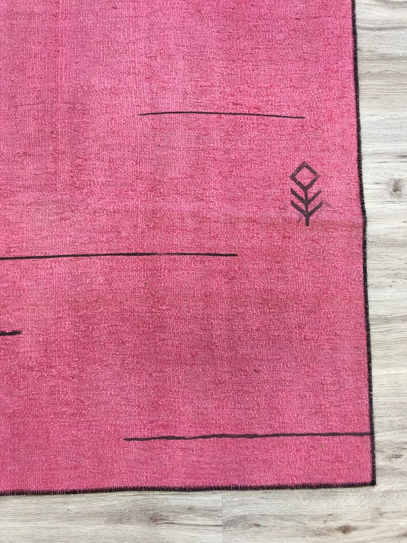 K&ouml;nnte beinhalten: Ein rosa Teppich mit einem schwarzen geometrischen Muster. Der Teppich hat einen Fransenrand.