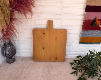 May include: A rectangular wooden chopping board with a central handle at the top. The board has a light brown finish with visible wood grain patterns and several dark circular knots distributed across the flat surface of the wood.