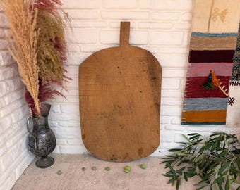May include: A large, weathered wooden cutting board with a rectangular handle at the top. The board has a rounded bottom edge and a natural, light brown wood grain finish with visible signs of wear, including small dark spots and surface texture.